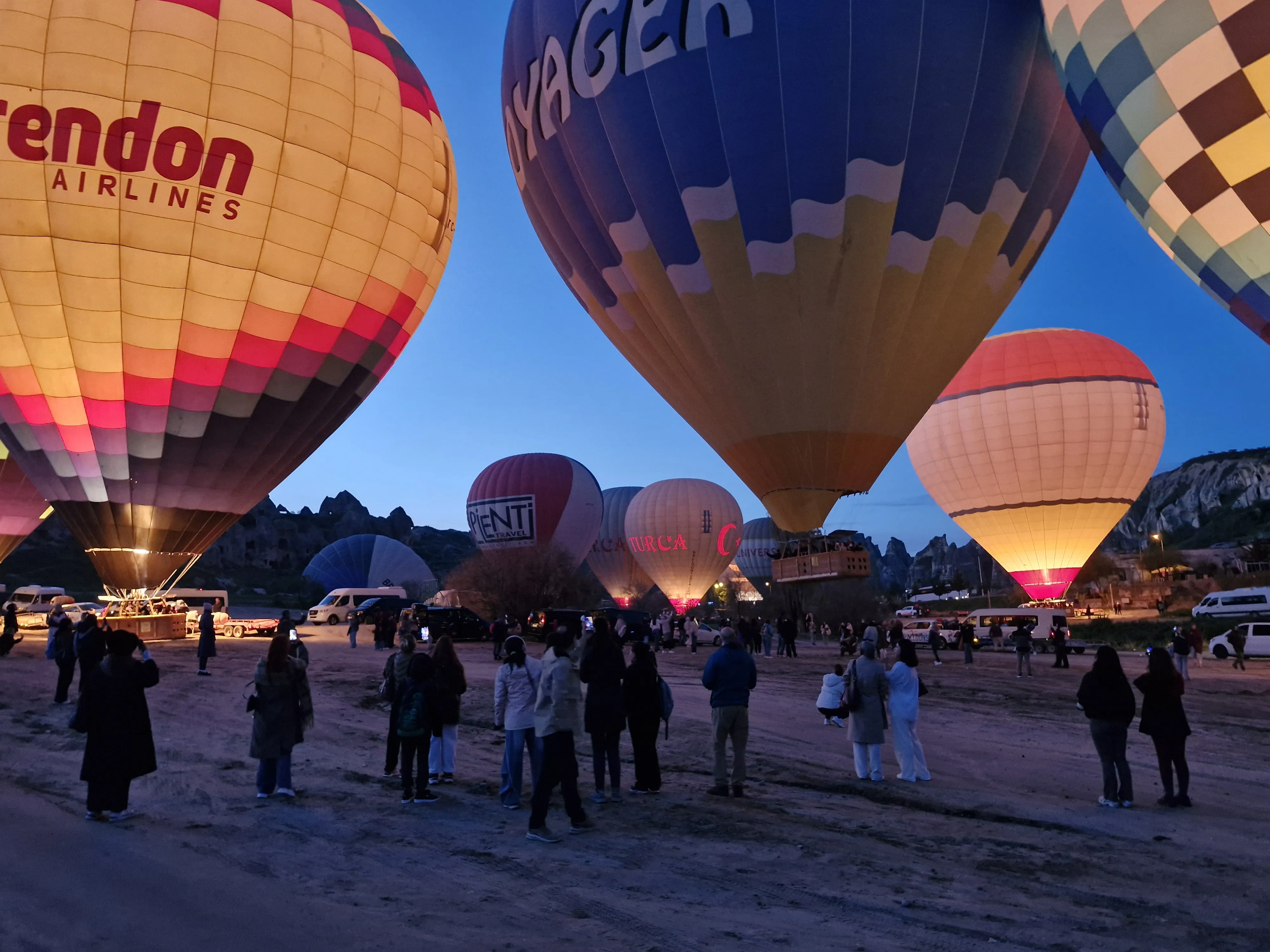 Hot air balloons preparing for sunrise takeoff in Cappadocia as visitors gather at the launch site.